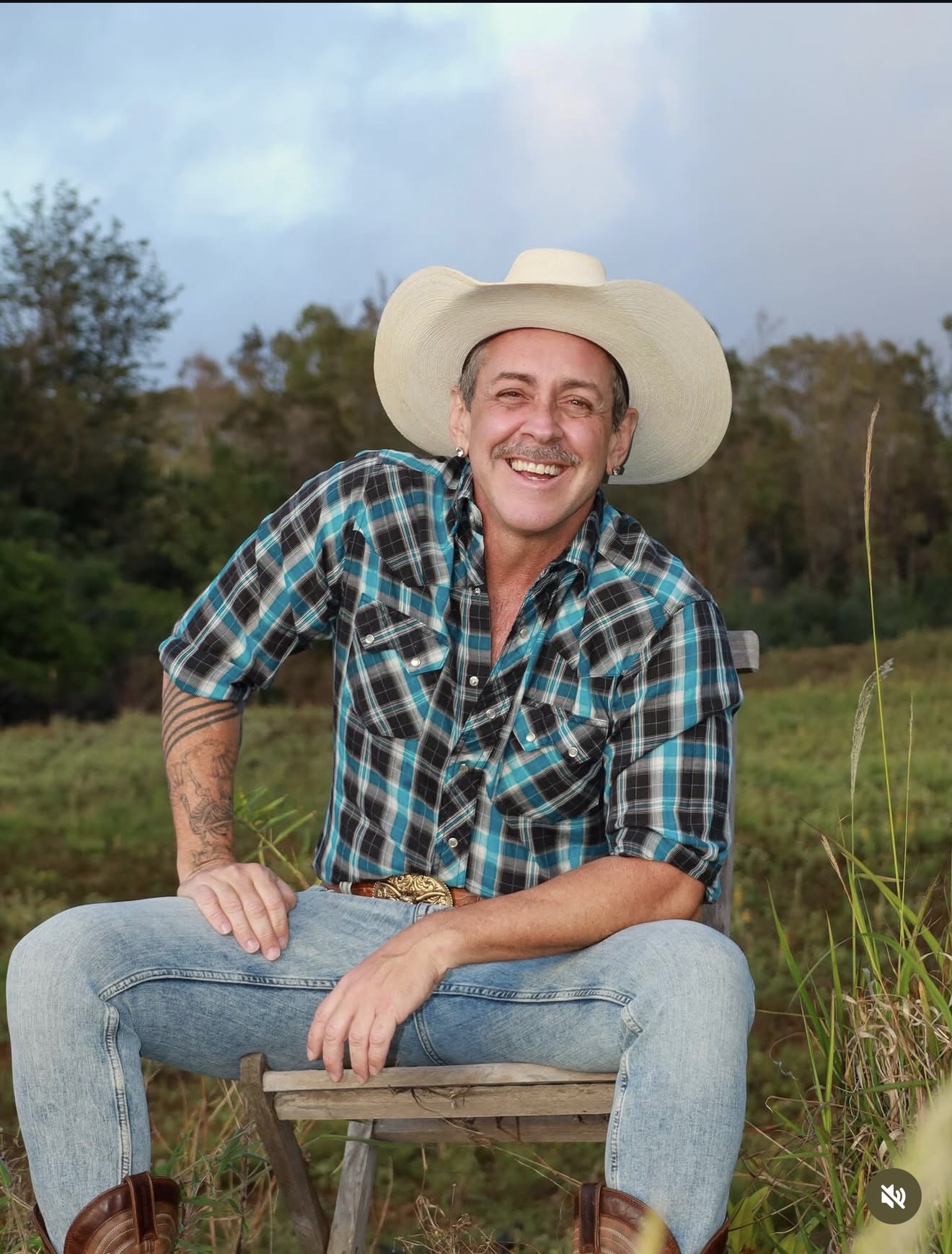 Malcolm Phoenix McCloud smiling in a cowboy hat and plaid shirt, sitting in a field at golden hour
